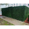Image of Artificial Boxwood Hedge Plant Wall Screening Panels serving as a privacy fence in an outdoor setting on a concrete walkway. In the background, a multi-story hotel or apartment building and several palm trees are visible