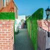 Image of A narrow outdoor walkway with Artificial Boxwood Hedge Plant Wall Screening Panels and brick pillars on one side, artificial turf underfoot, and a brick building with a staircase on the other, leading toward a distant reception area