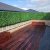 Image of A modern backyard with a reddish brown wooden deck, tall Artificial Jasmine Hedge Plant Wall Screen Panels, matching storage benches, and neighboring rooftops in the background
