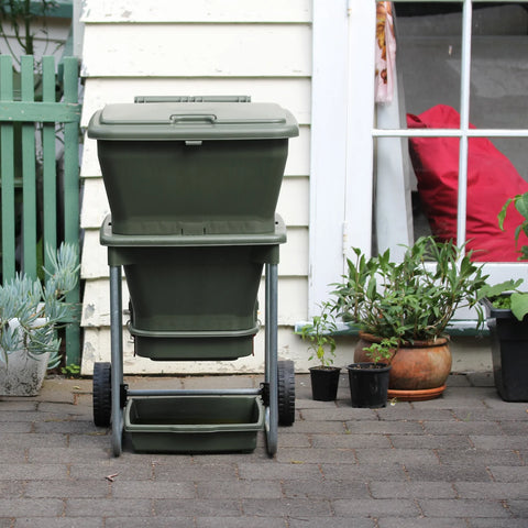 Green hungry bin on wheels outside a house with plants around

