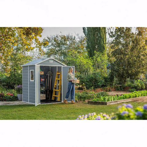 Person standing outside a garden shed in a garden setting with trees and plants.