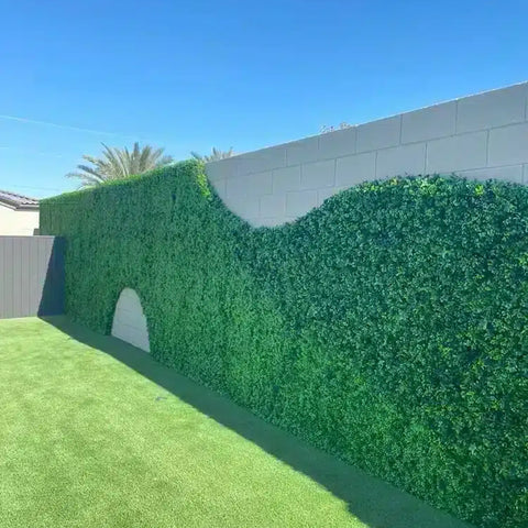 A modern backyard featuring wavy topped Artificial Flowering Buxus Hedge Plant Panels on an off-white wall, vibrant green turf, and palm fronds peeking above under a sunny sky