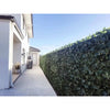 Image of Artificial Peach Leaf Ivy Hedge Screen installed on a backyard fence, set beside a cement pathway with a large potted plant, windows, and a backyard door in view