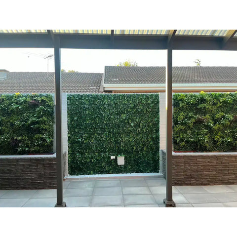 The center features Artificial Ivy Leaf Hedge Panels while the flanking panels showcase varied vertical gardens with mixed artificial plant textures and species set above low decorative stacked stone planter boxes in front of the paved outdoor living space