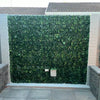 Image of An image of a large Artificial Ivy Leaf Hedge Panel installed on an outdoor feature wall, framed by light gray brick pillars and contrasting with gray square tiles below with precise cuts around a small utility box