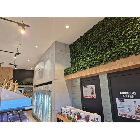 A modern commercial space featuring the Artificial Ivy Leaf Hedge Panel on a light wood base adding a contrast to the store's industrial design which includes gray concrete style panels, air condition, commercial refrigerators, modern track lighting, product displays and signage