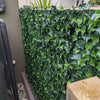 Image of Close up view of the Artificial Ivy Leaf Hedge Panel installed in a narrow outdoor passage. The ground is covered with gravel with a large utility pipe on the left and a thick wooden fence post is visible in the immediate foreground